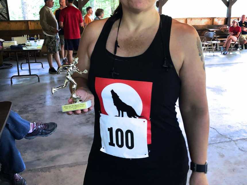 A woman wearing a black tanktop holds a trophy after running a race. On her shirt is a wolf howling.