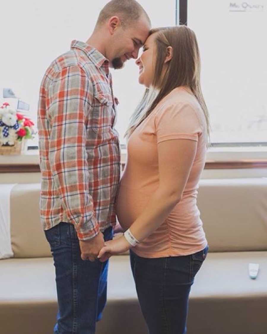 A man and his pregnant wife hold hands and look at each others. She is wearing an orange shirt and he is wearing plaid.