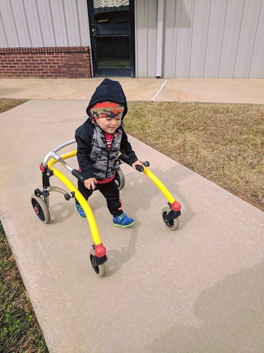 A toddler wearing two jackets uses a yellow walker to walk along a sidewalk.