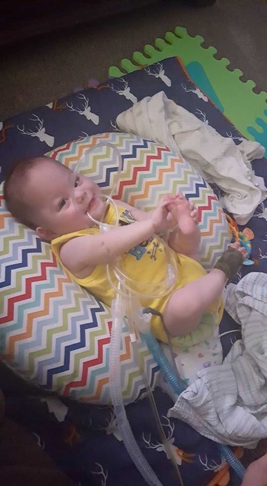 A little boy in a yellow shirt bites on his oxygen tube while lying reclined on a rainbow-striped pillow.