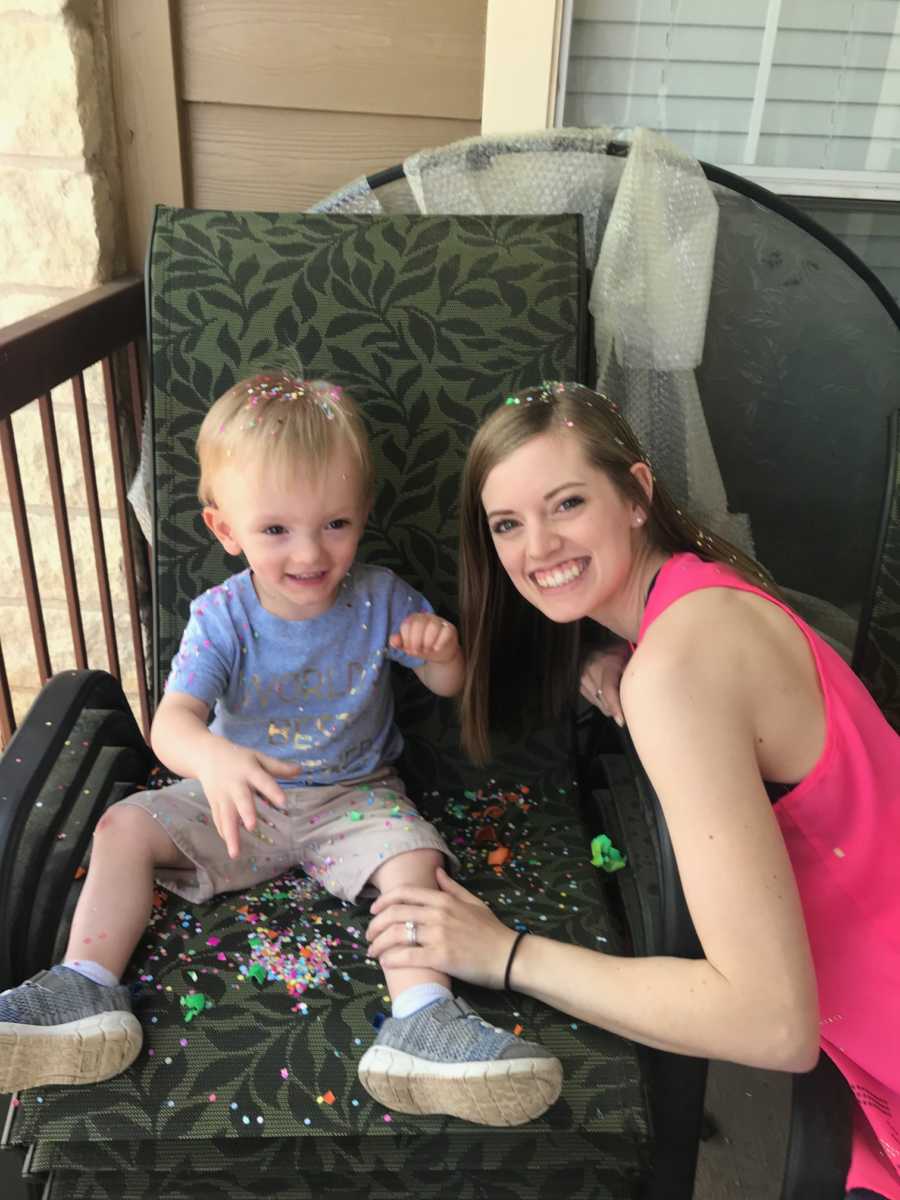 A mom crouches next to her young son, who sits on a chair in shorts and a blue T-shirt covered in confetti.
