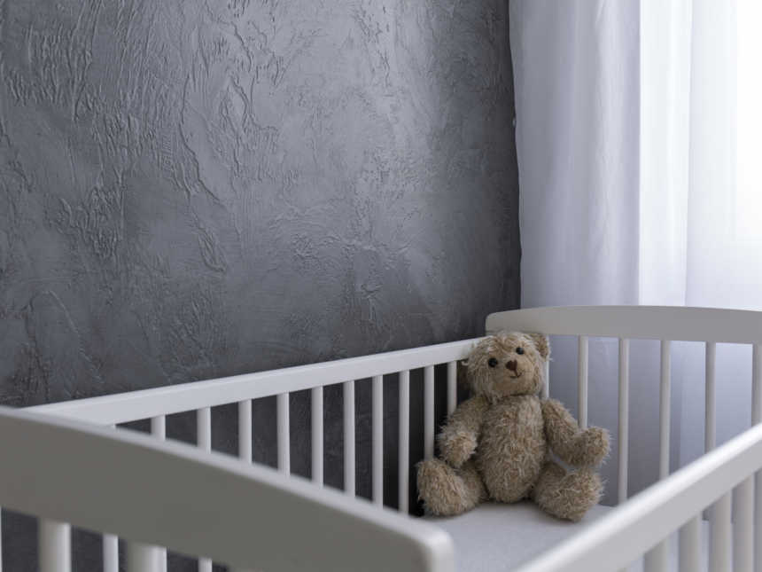 A white crib in a grey room near white curtains with a brown teddy bear
