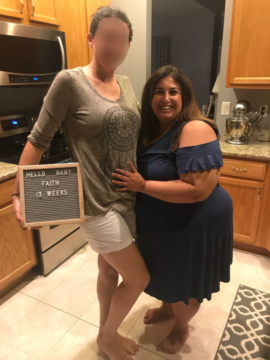 Two women stand together in a kitchen with one having a hand on the other's stomach