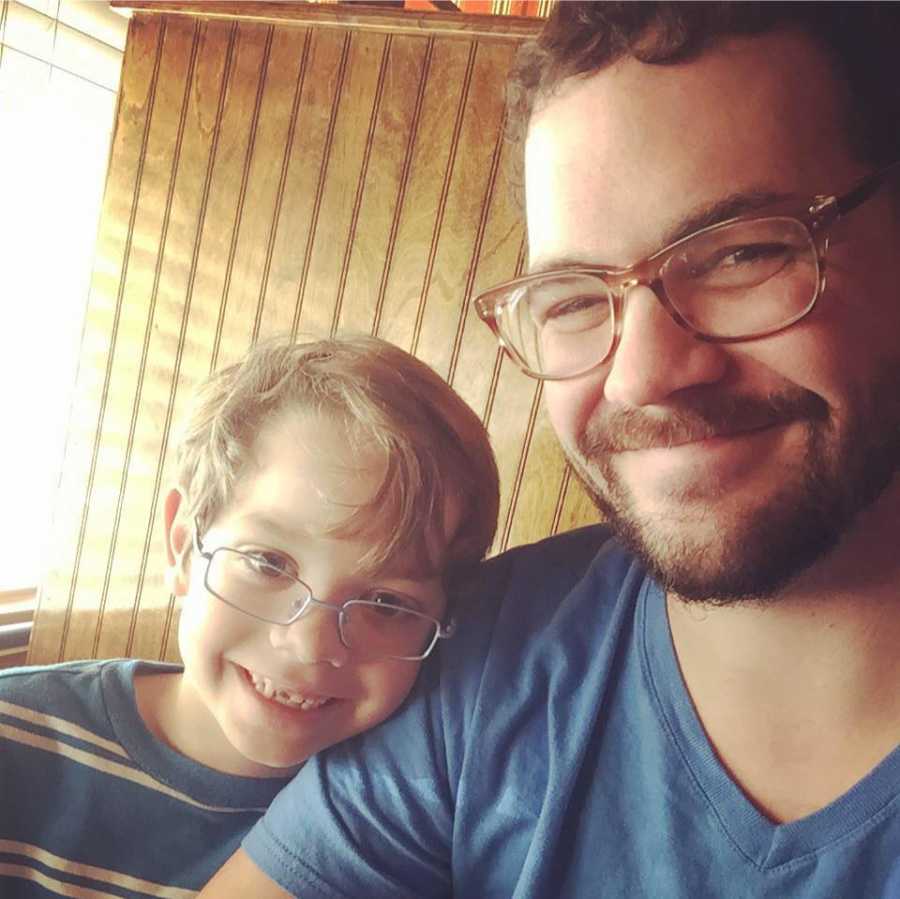 A dad in a blue shirt and glasses sits with his son, who is also wearing glasses and a blue shirt.