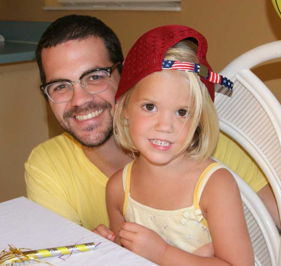 A dad in a yellow shirt and glasses kneels next to his daughter in a yellow shirt and American flag ballcap.