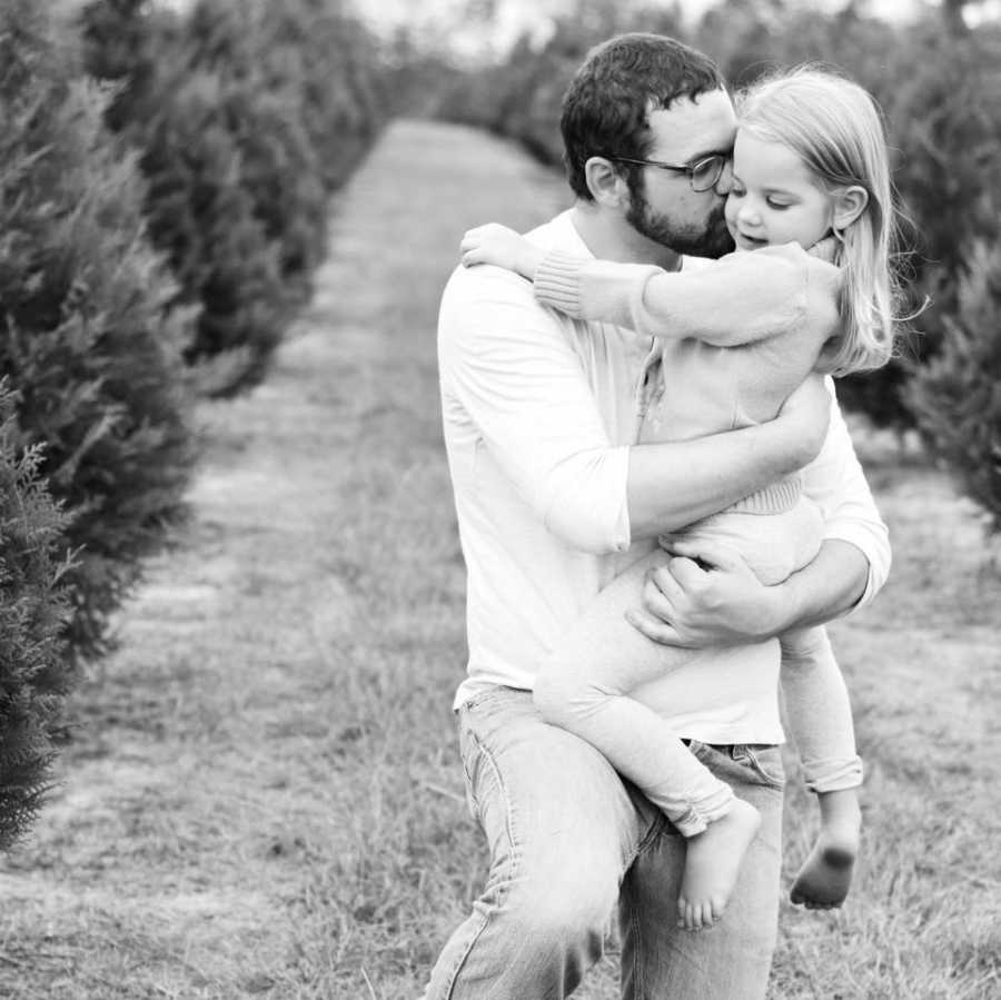 A dad holds his daughter in his arms and plants a kiss on her cheek while standing outside between rows of trees.