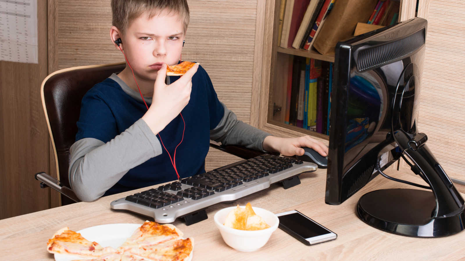young boy eats pizza while using computer and looking irritated