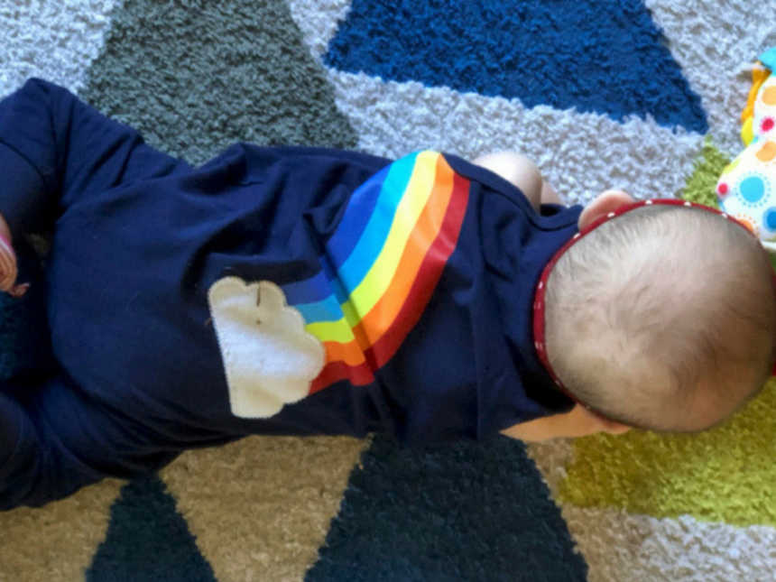 A baby wearing a blue onesie with a rainbow on it lying on a rug with blue and gray triangles