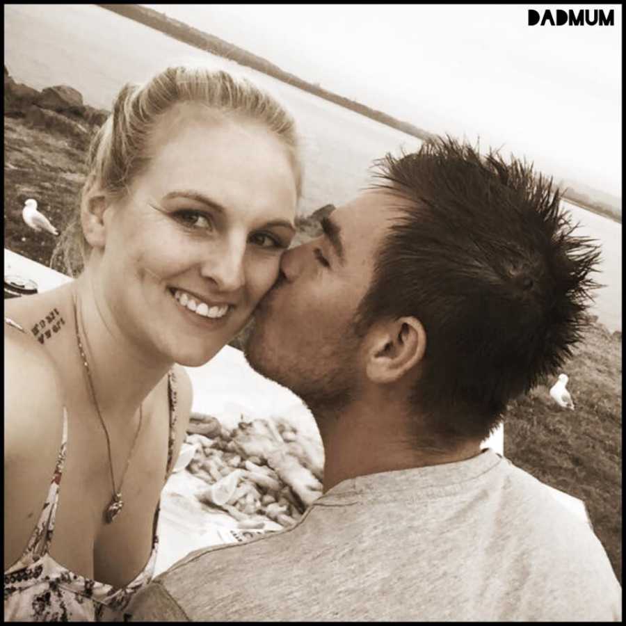 Woman smiles for a selfie while her partner kisses her on the cheek during a beach picnic