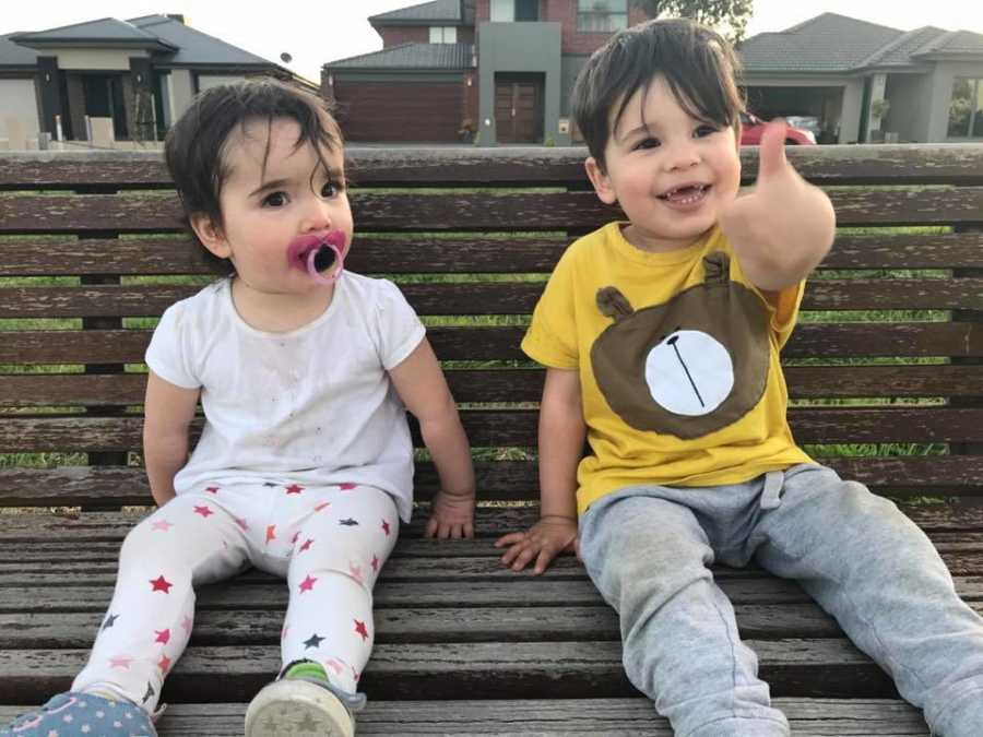 Siblings sit on park bench together while older brother gives the camera a thumbs-up