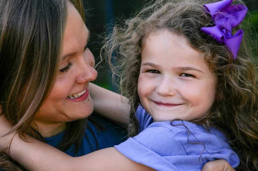 Mom smiles at daughter while she smiles for the camera in purple dress and bow