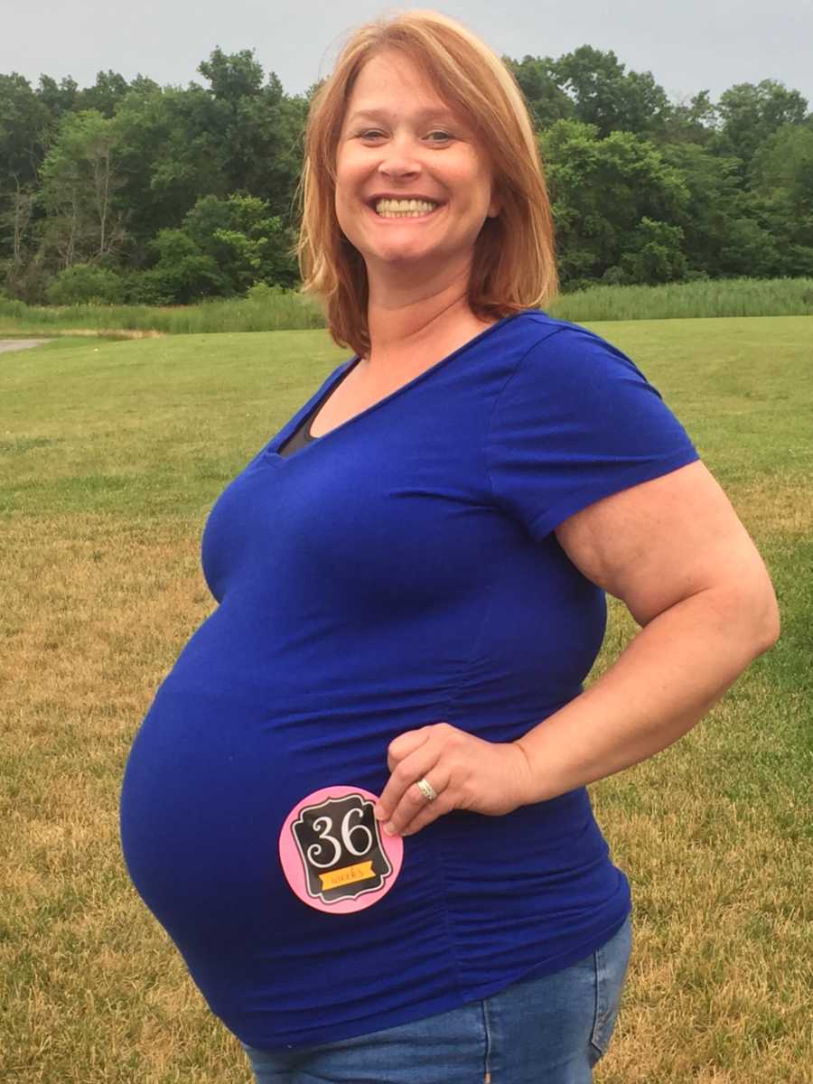 Surrogate for her daughter smiles for a photo while holding a sign that says she's 36 weeks pregnant