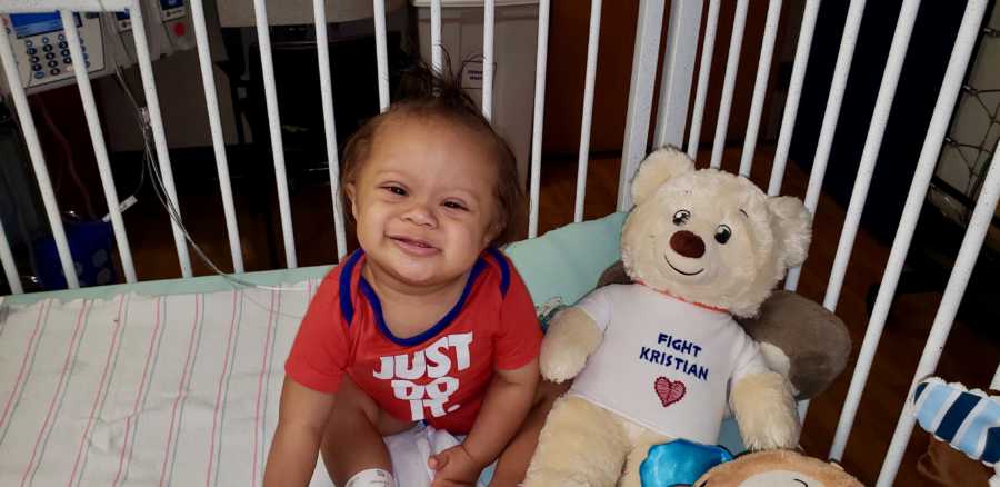Little boy with blood disorder smiles for a photo while sitting next to a bear with a shirt that reads "Fight Kristian"