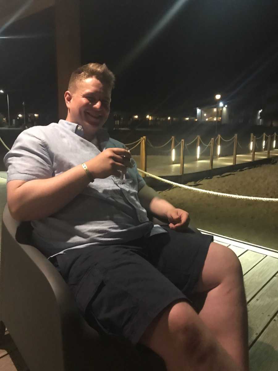 Man on vacation smiles for a photo while holding a drink and sitting on a beach at night