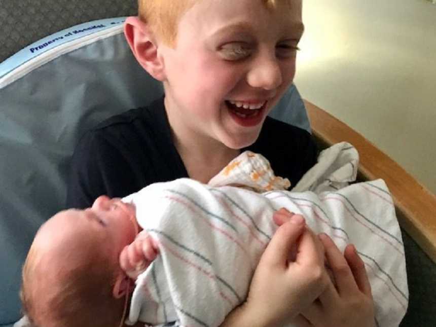 Big brother laughs while holding his swaddled baby brother