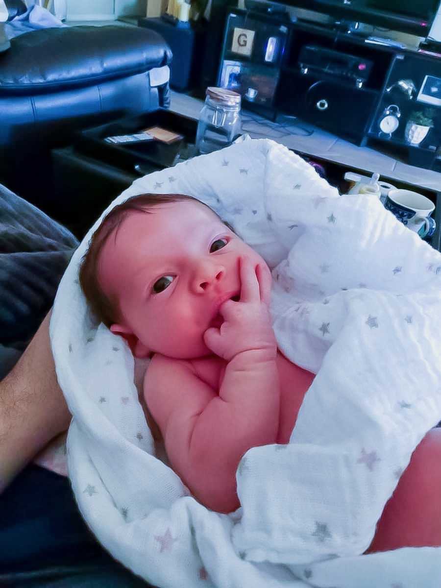 Mom snaps a photo of newborn chewing on her fingers while swaddled in mom's lap