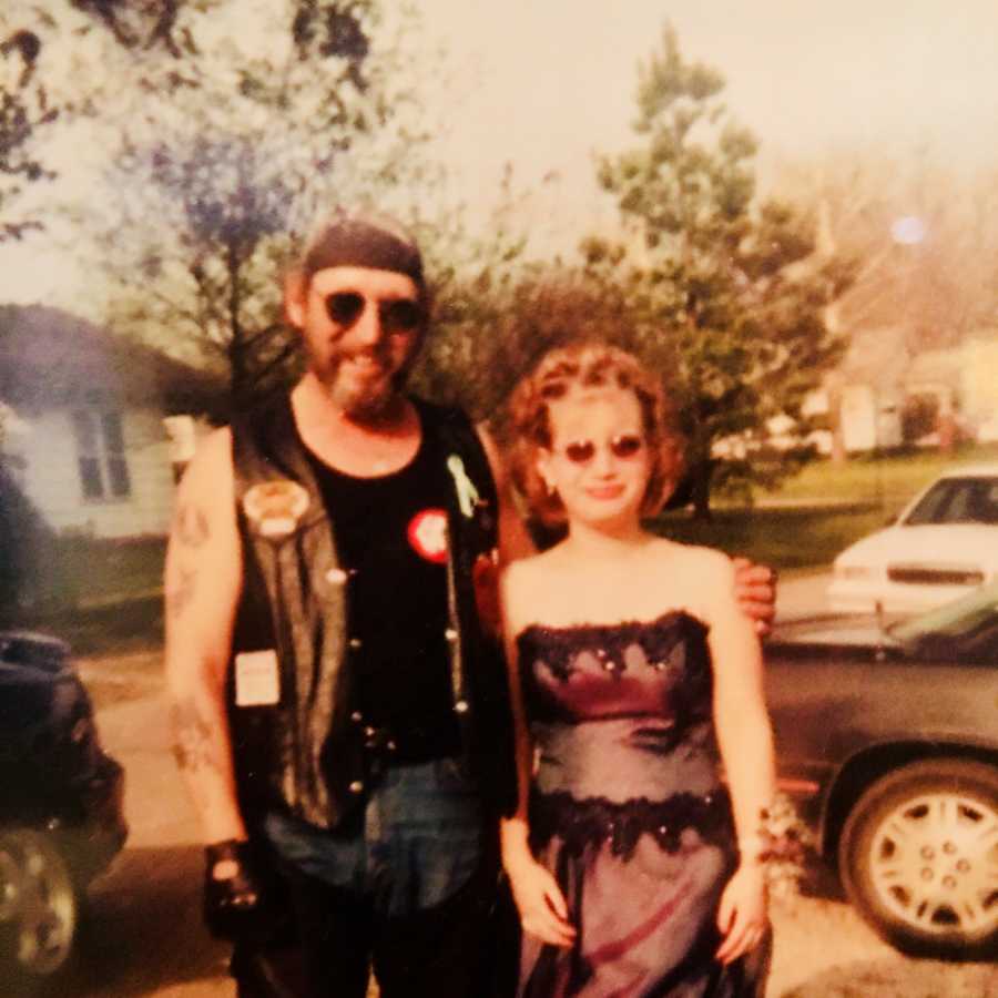 Teen girl stands in prom dress next to biker dad for a photo