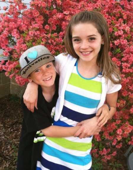 Mom takes a photo of her daughter and son while they stand in front of floral bush