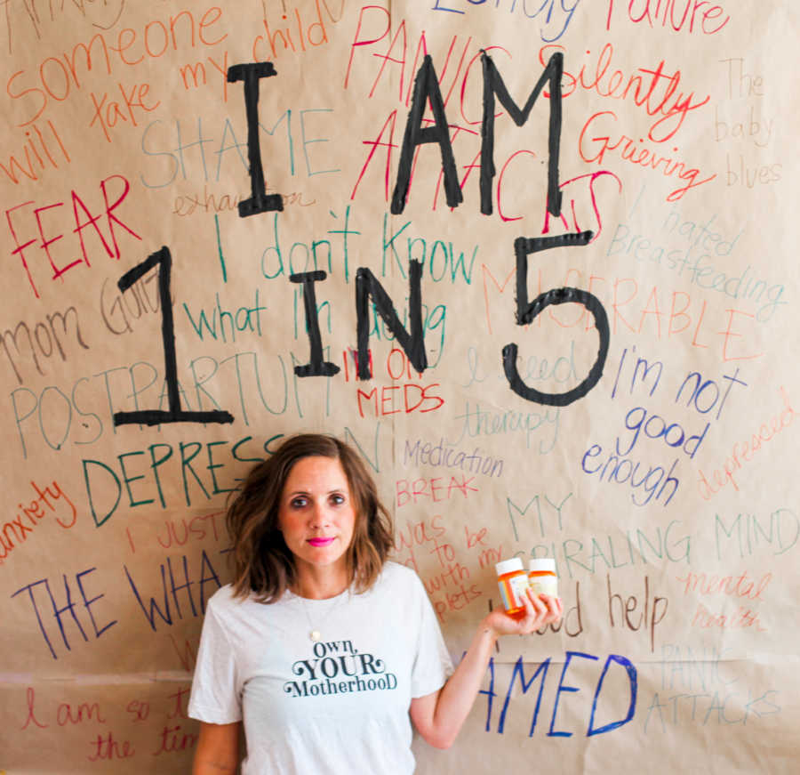 Woman with postpartum anxiety holds her anxiety medication while standing in front of a poster that reads "I am 1 in 5"