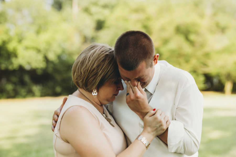 Groom stands holding his face as he cries as mother with breast cancer holds him