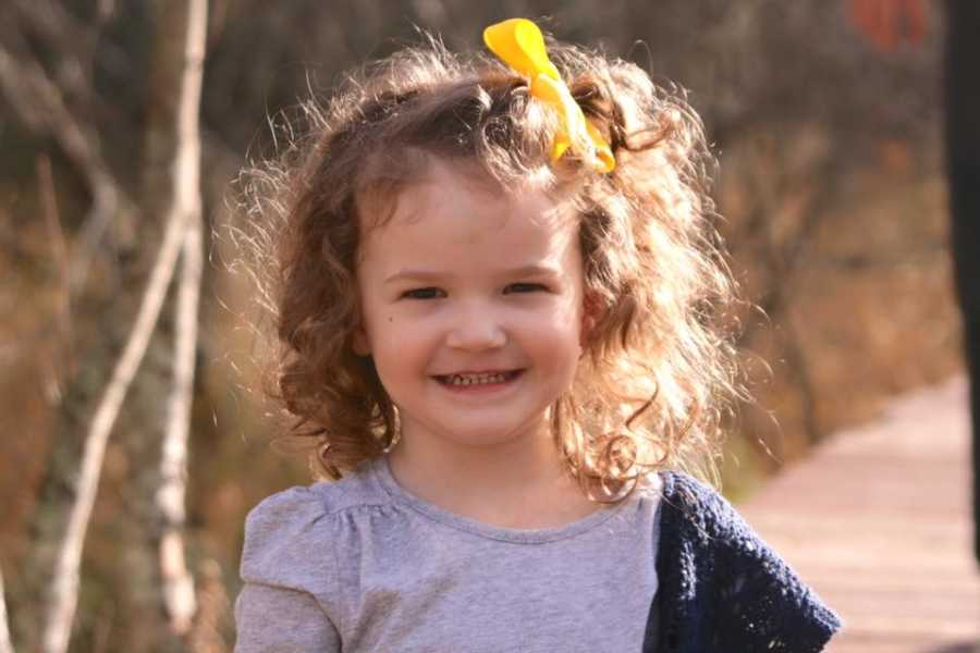Young girl standing and smiling with yellow bow whose mother is nervous for her to start school