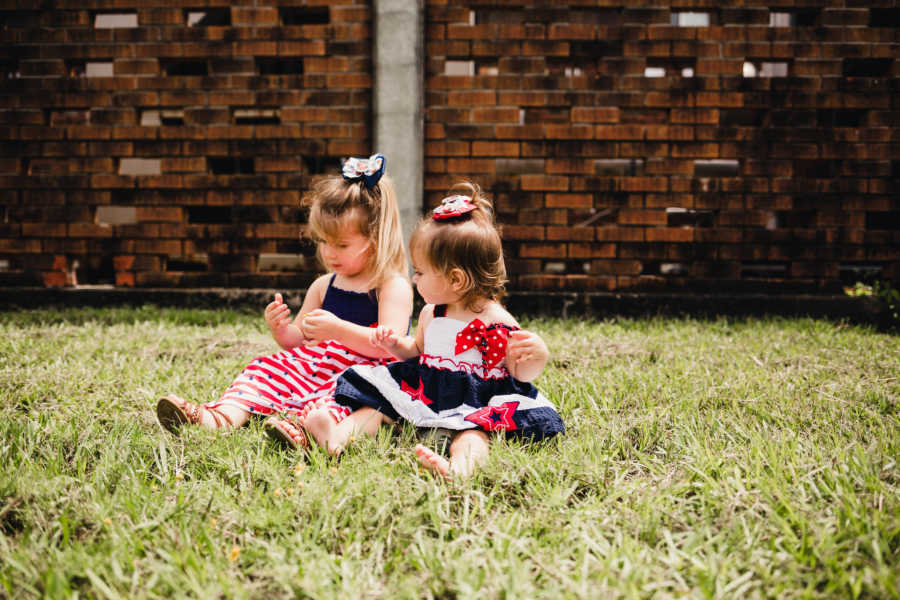 Two little girls sit in grass wearing red white and blue awaiting their soldier father's return