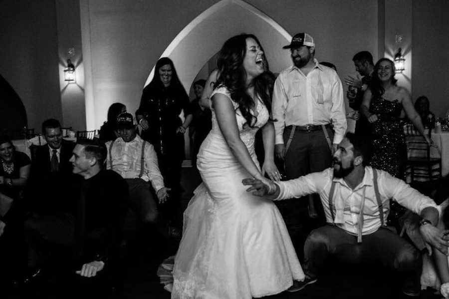 Bride laughs while holding husband's hand who is crouching to ground with people watching
