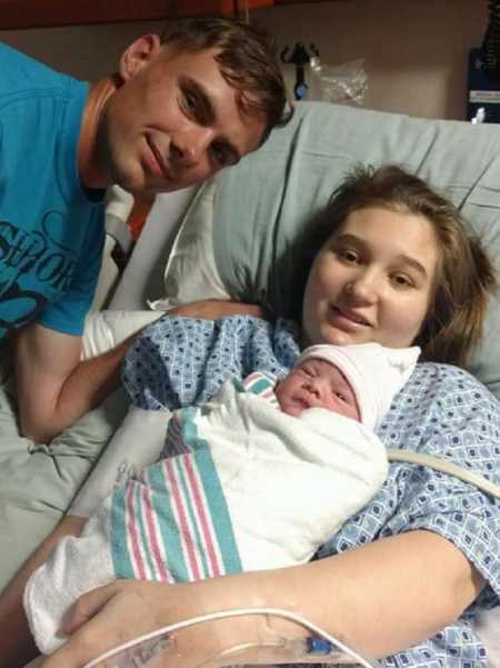 Mother lays in hospital bed smiling with newborn daughter on her chest beside her husband