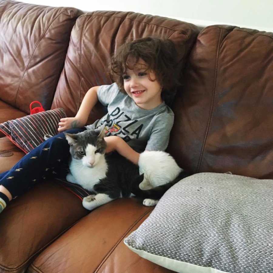 Young boy whose mother is proud of him for using hair dryer sits on couch smiling with cat