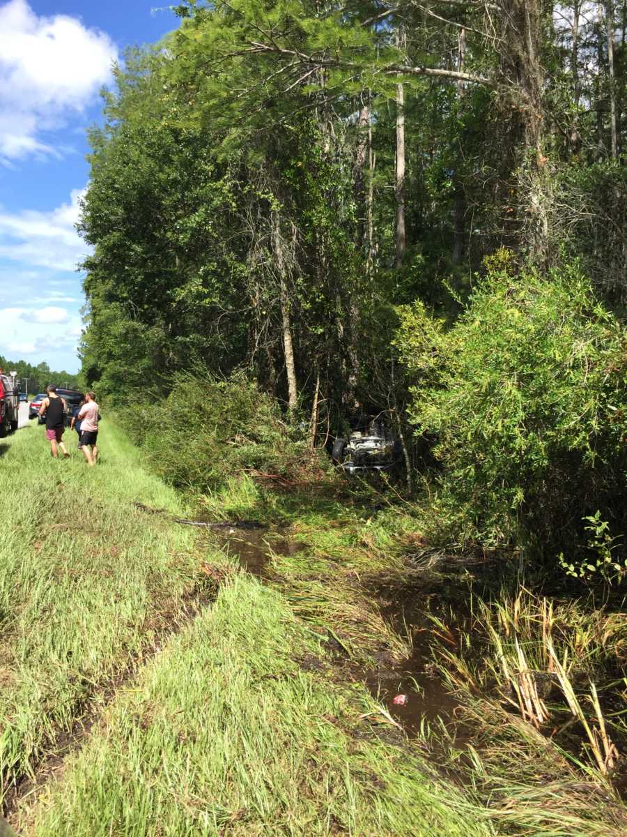 Car flipped over in ditch with people standing on side of road 