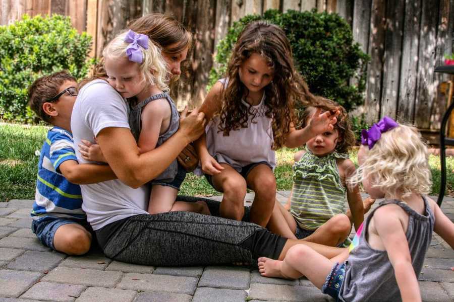 Woman sits on ground outside with twin daughters, little boy who she has custody of and two other little girls