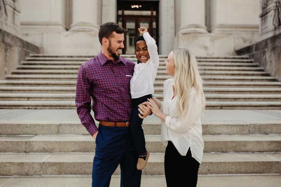 Husband and wife hold adopted son who raises an arm in air on steps outside of adoption court