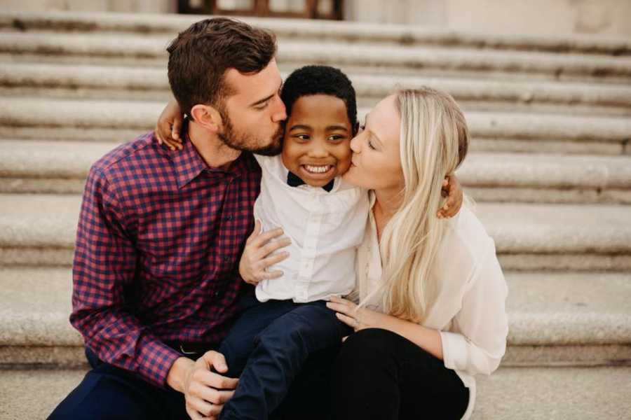 Husband and wife sit on steps outside of adoption court kissing the cheeks of their adopted son