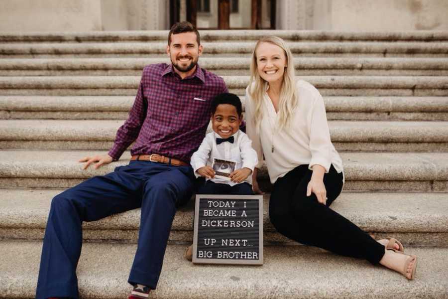 Husband and wife sit on steps of adoption court with adopted son who holds ultrasound for new sibling