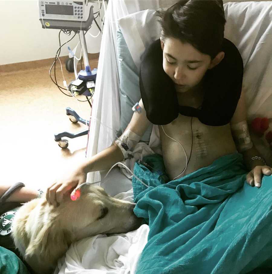 12 year old with heart condition and scar on chest sits up in hospital bed petting dog beside him