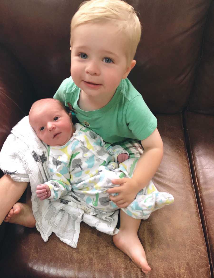 Toddler boy holds newborn little brother on a brown leather couch