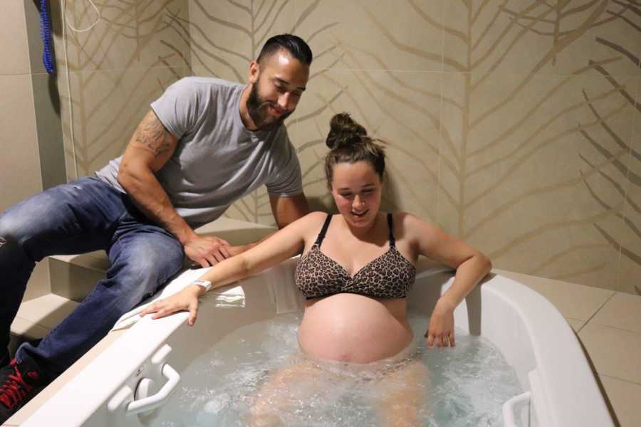 Woman sits in birthing tub at home in a cheetah print bra while her husband sits on the tub beside her