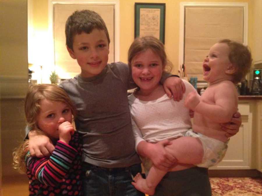 Three young kids smile arm in arm while one holds baby whose mother needs help taking care of her