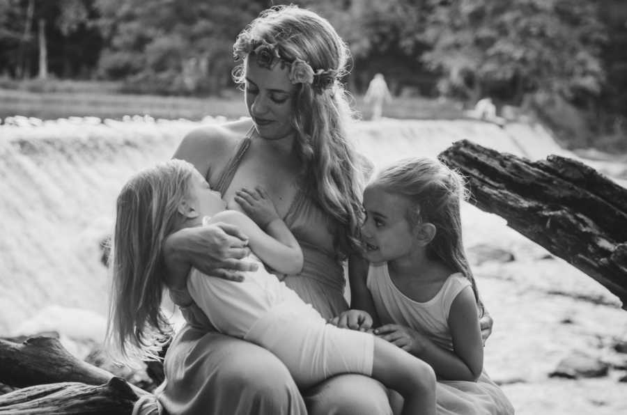 Mother sits breastfeeding 3 year old beside her oldest daughter in front of roaring river