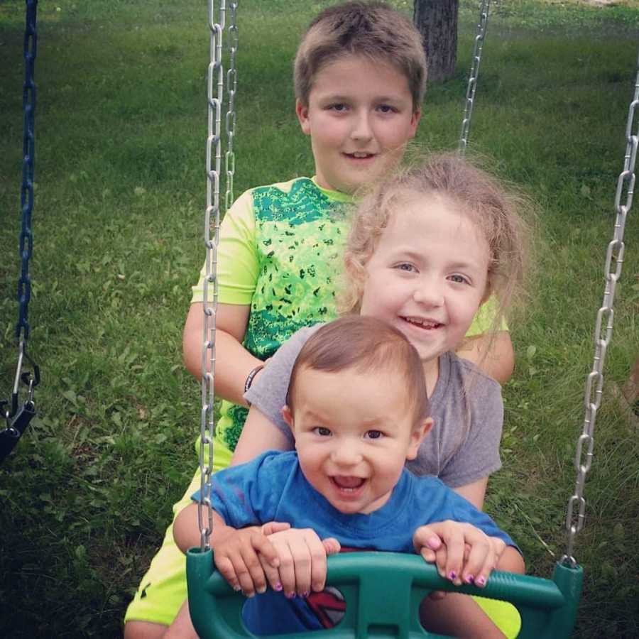 Oldest brother sits with younger sister and brother on swing whose mother had miscarriage with 4th sibling