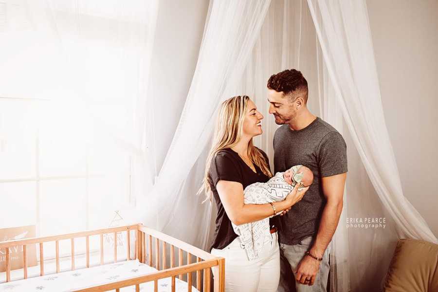 Husband and wife look into each others eyes while holding adopted newborn next to crib