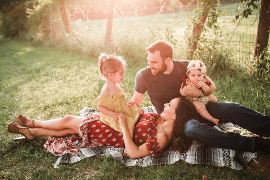 Woman who talks about reality of parenting lays down with daughter sitting on her next to husband and other daughter