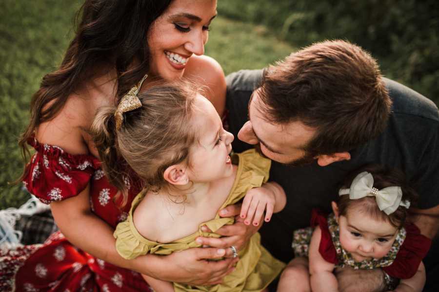 Mother who speaks on reality of parenting sits with daughter in lap looking at father who is holding younger daughter