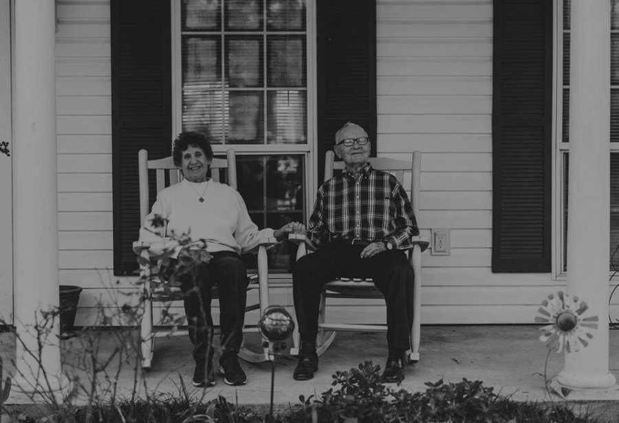 Elderly woman with dialysis sits smiling in rocking chair beside husband outside their home