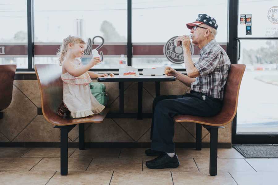 Toddler sits holding milk bottle in booth across from grandpa who is drinking milk 