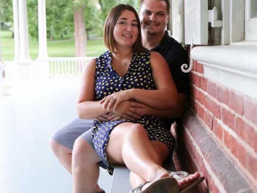 Husband smiles sitting on bench beside pregnant wife with his arms wrapped around her