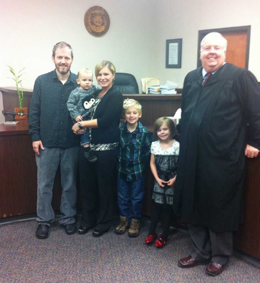 Husband and wife stand with their adopted baby beside two other adopted children and judge in adoption court