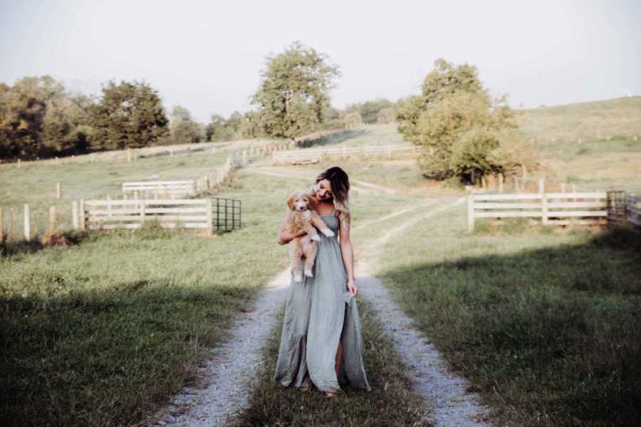 Recently single dog mom walks along dirt path on farm holding puppy