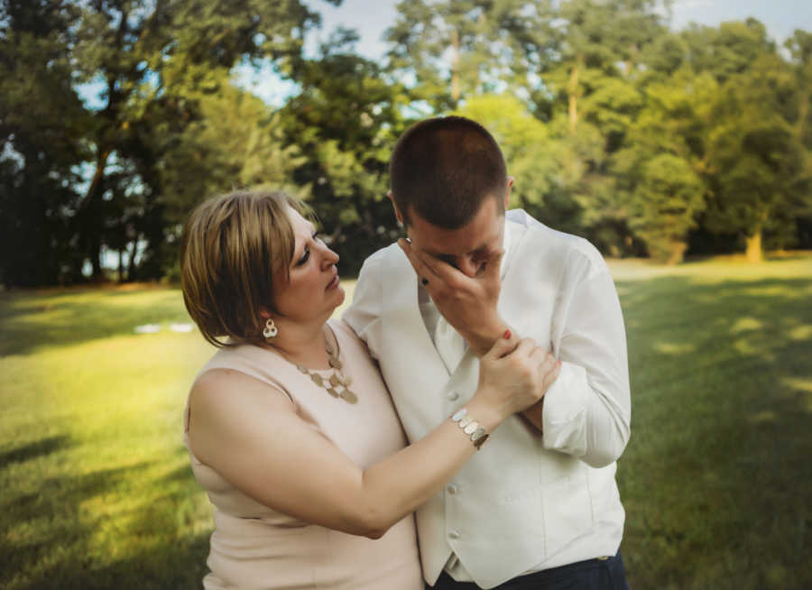 Groom crying with hand to his face while mother with breast cancer holds him