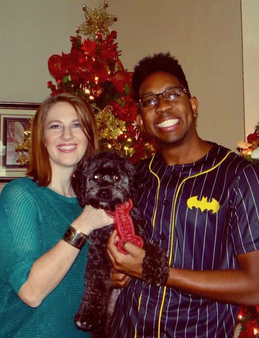 Husband and wife smiling holding dog in front of Christmas tree